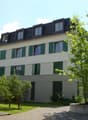 3 story apartment building, white walls, green shutters, gravel driveway, tree, plants, barrel