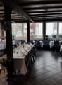 A restaurant interior with wooden beams, large windows, dark tiled floors, and tables with white tablecloths and neatly arranged wine glasses and napkins.