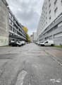 A street with parked cars along the sides, two tall buildings, concrete sidewalk, a few trees, and cloudy sky