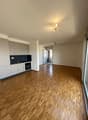 The image shows a spacious open-plan apartment with a modern kitchen and living area. The apartment features hardwood flooring throughout, white walls, and recessed lighting. There is a sliding glass door leading to what appears to be a balcony or terrace