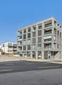A modern, multi-story commercial or office building with a rectangular, glass-and-concrete facade. The building is situated on a street corner with a road and sidewalk visible in the foreground.