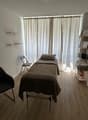 Simple bedroom setup with a bed, a foldable chair, and a shelf with certificates, located in a room with white walls, wooden floor, and a large window with curtains.