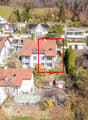 Aerial view of a residential area with several houses, one of which is highlighted with a red square. The highlighted house has a brown roof and white walls. The houses are surrounded by trees and greenery. The area is on a slope, with some houses having