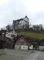Several houses on hill, larger one at top, brown wooden houses below, small town with cars
