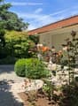 Garden with gravel path, various plants and flowers, brick building with garage, and a white chair.