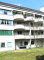 3-story apartment building with white exterior, balconies on each floor, and large windows. The building has a green roof and is surrounded by a grassy area with trees.