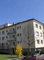 4 story apartment building, beige exterior, multiple balconies, windows, green bushes, trees, cars parked
