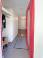 The entryway of a building with red door, tiled floors, a small carpet, a closet with clothes and shoes visible, a wooden shelf with shoes, and a hallway leading to a white door. The hallway is illuminated by a light fixture on the ceiling. The entire int