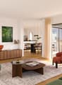 Spacious open-plan living room with large windows, hardwood floors, modern furniture including a wooden sideboard, coffee table, and armchairs in various colors. The room has access to a balcony or terrace through sliding glass doors.