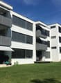Modern white apartment building with multiple balconies, well-maintained garden, and outdoor seating area.
