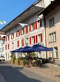 This image shows a street view of a quaint European town. The buildings have traditional architecture with red shutters and wooden beams. There are several restaurants and shops along the street, with outdoor seating areas under blue umbrellas. Bicycles a