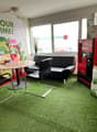Small room with round table, two black chairs, a red sofa, a Coca Cola machine, a poster on the wall, a potted plant on the windowsill, and artificial grass flooring.