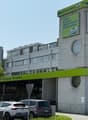 A multi-story commercial building with a Steinmann cafe and restaurant on the ground floor. The building has a large green and white sign advertising their cafe, lunch, take away, and chocolate offerings. There are several cars parked in front of the buil