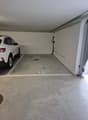 The image shows a garage space with a white SUV parked inside. The floor is concrete and there is a number '15' painted on the floor, indicating this is parking spot number 15. The walls are plain white and there are some utility pipes or wires visible on