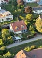 House with red roof, car parked in garage, surrounded by trees and grass, aerial view