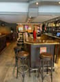 bar counter, stools, wooden flooring, wooden bar, bartop, shelves with alcohol bottles, gaming machines, plants
