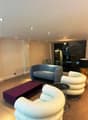 Spacious living room with modern furniture, including a blue sofa, white and beige armchairs, and a purple coffee table on wheels. The room has recessed lighting in the ceiling and a stone accent wall with a fireplace.