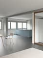 Spacious open-plan living area with a kitchen, dining table, and living room. Large windows provide ample natural light. The kitchen features blue cabinets and a minimalist design. The dining table and chairs are made of wood, complementing the overall mo