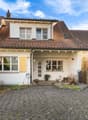 Detached 2-story house, red tiled roof, white walls, yellow shutters, a driveway, fenced front yard, several plants in front