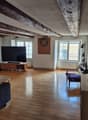 Spacious living room with hardwood floors, exposed wooden beams on the ceiling, a large TV mounted on the wall, a sofa, and a coffee table. There is also a dart board on the wall and a potted plant in the corner.