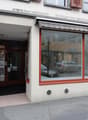 The image shows the exterior of a commercial building with a storefront. The building has large windows with red frames, and the entrance is through a doorway. There are cars parked on the street in front of the building.