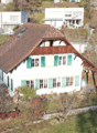 White house with brown roof, green shutters, surrounded by trees, elevated on the slope, garden