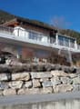 Two-story house, white exterior, balcony with railings, stone retaining wall, mountains in the background.