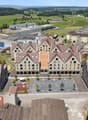 The image shows an aerial view of a town with a mix of residential and commercial buildings. The town has a central square or plaza surrounded by multi-story buildings with red tile roofs. There are also some industrial or warehouse buildings in the backg