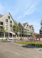 The image shows a residential area with several multi-story apartment buildings. The buildings have a modern, uniform architectural style with gabled roofs and large windows. There is a public plaza or square in the foreground with benches, bike racks, an