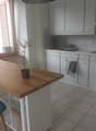 Open kitchen with a wooden island, white cabinets, white tiled floor, a window with shutters, a sink with faucet, potted plant, a kettle, toaster, and a cooking stove.
