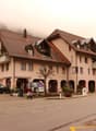 A row of multi-story residential buildings with red tile roofs, located in a mountainous area with fog in the background. The buildings have shops or businesses on the ground floor and apartments above. There are parked cars in front of the buildings.