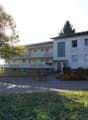 2 story white building, multiple windows, balcony, parking lot, trees, plants, green car