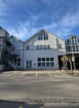 A multi-story commercial or office building with a modern, glass-heavy architectural design. The building has a large central structure with several wings and annexes. The exterior is primarily composed of light-colored walls and large windows. There is a