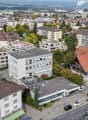 Aerial view of several multi-story residential buildings, green spaces, and trees. Several vehicles parked in the area.