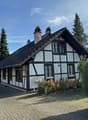 Detached house, black roof, white and brown walls, chimneys, car parked in front, brick driveway, garden, trees