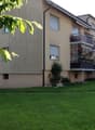 Apartment building, beige walls, three floors, multiple balconies, green lawn, trees, plants