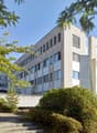 Office building, light grey facade, many windows, concrete base, multiple floors, green shrubs, trees, steps to the entrance.