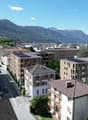 Aerial view of a town with several buildings, mountains in the background, solar panels on some buildings, cars parked on the street, trees and greenery