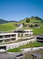 Modern architectural design, multi-story building, white exterior, large windows, solar panels on the roofs, located on a hillside with green surroundings, stone walls, and gardens.