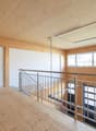 The image shows a large open space with a wooden ceiling and walls. There is a metal railing along the second floor balcony, and the floor is made of wood planks. The room has large windows that provide natural light. The overall design appears to be mode