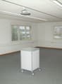 Large open room with white walls, ceiling-mounted projector, and multiple windows providing natural light. The room has a carpeted floor and a mobile cart or cabinet in the center.