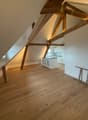 The image shows an attic or loft space with exposed wooden beams and trusses. The floor is made of hardwood planks, and there is a kitchen area visible in the background. The space has a cozy and rustic feel with the wooden elements.
