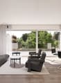 Spacious living room with large windows overlooking a lush, green outdoor area. The room features a grey sofa, black armchairs, a white dining table with chairs, and a potted plant. The walls are white, and the floor is made of wood. There is also a frame