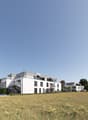 Modern, multi-story apartment building with white exterior, large windows, and balconies. The building is situated on a grassy field with yellow flowers.