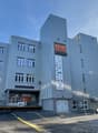 A multi-story commercial building with a gray metal facade and large windows. The building has the name 'Klubus' displayed prominently on the front. There are parking spaces in front of the building.