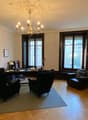 Wooden floor, wooden desk, three black chairs, one armchair, lamps, large windows, chandelier