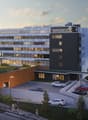 hotel with modern architecture, rooftop garden, many windows, parking area, multiple entrances, people walking around, various cars parked