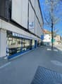 Commercial building with a shopfront, blue awning, and a sign reading 'Messer Wyss'. The building has a modern design with glass windows and a white wall. There is a parking sign and a tree in front of the building.