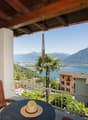 Wooden balcony with a scenic view of a lake surrounded by mountains and palm trees. The balcony has a wooden chair and a straw hat, suggesting a relaxing outdoor space.