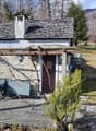 Stone house with chimney, brown door, stone pathway, wooden fence, grassy area, tree, plants, distant mountain